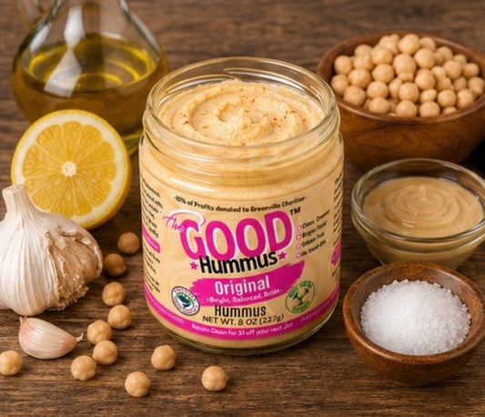 Jar of The Good Hummus on a wooden surface with ingredients like chickpeas, lemon, olive oil and garlic.