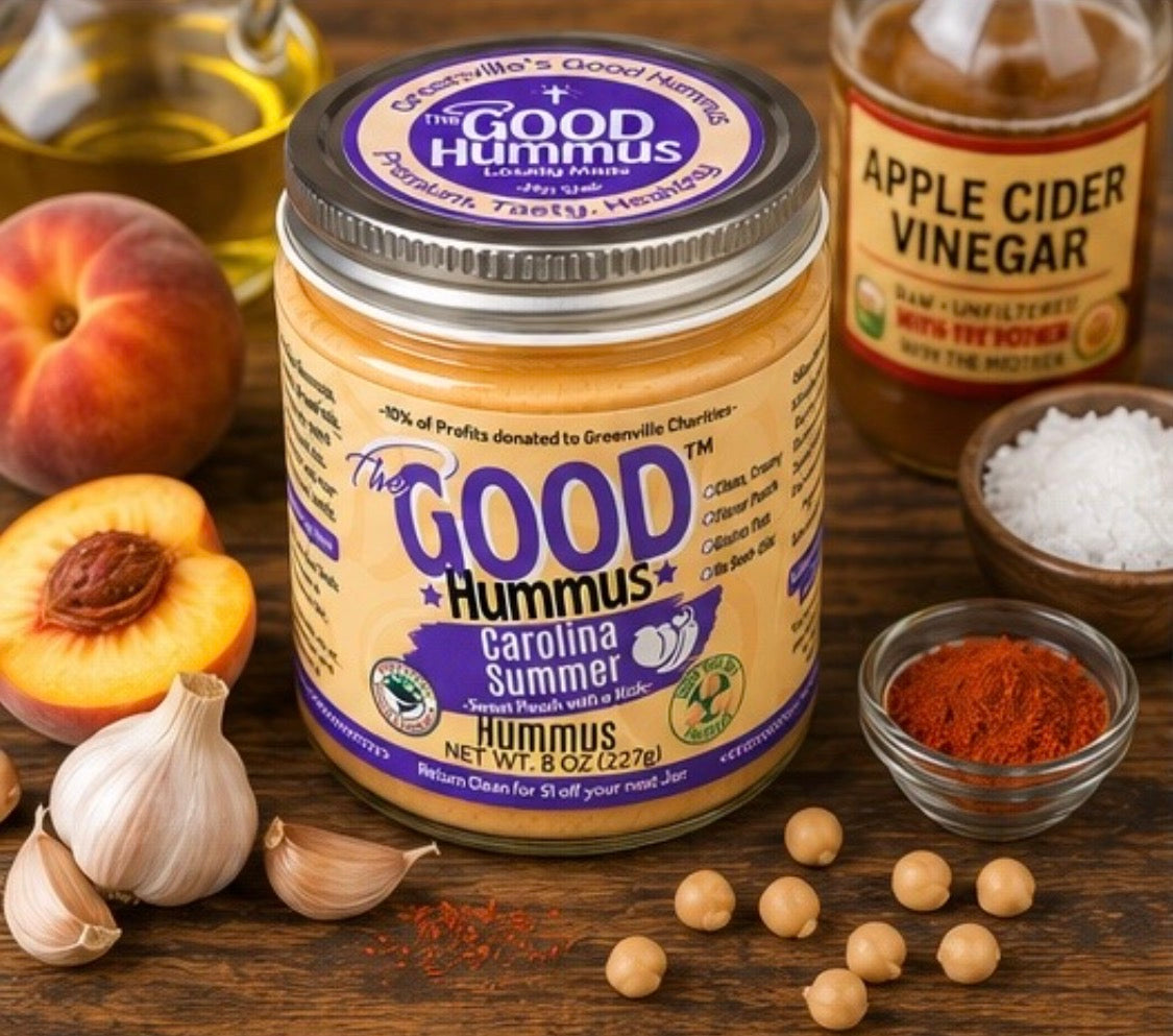 Jar of 'The Good Hummus Carolina Summer' on a wooden surface with ingredients like peaches, garlic, and spices.
